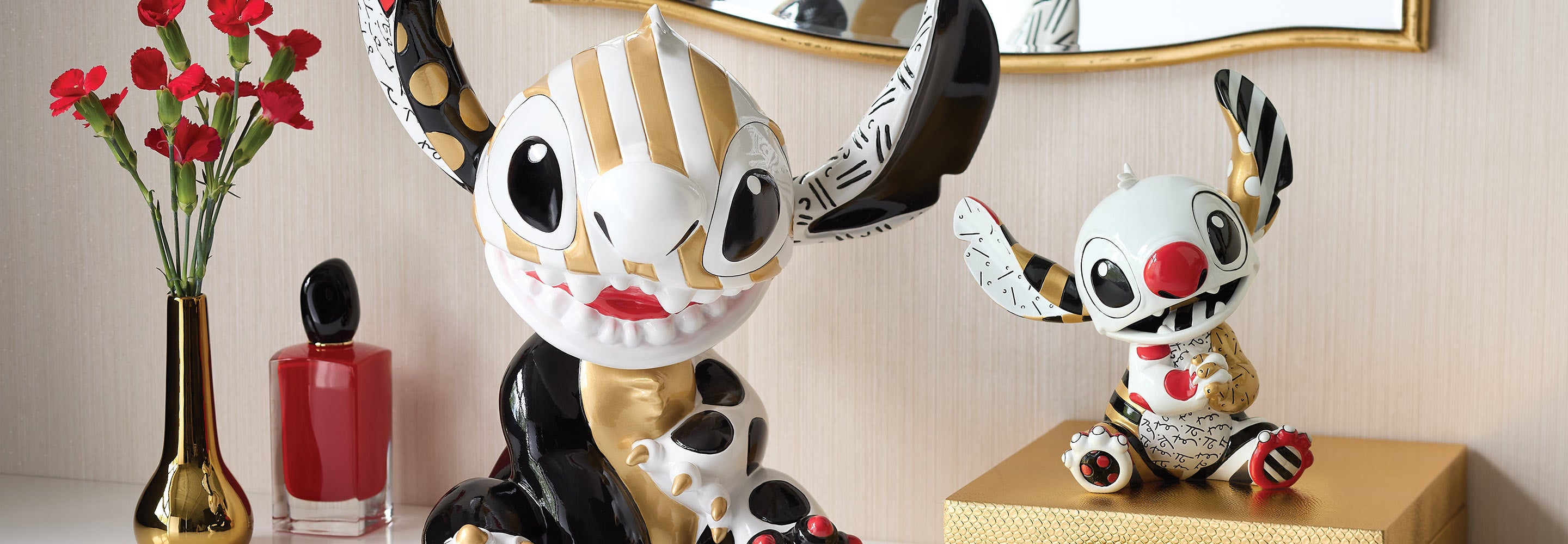 Two decorative figurines with large eyes and unique designs on a surface with a vase and glass of red liquid in the background.